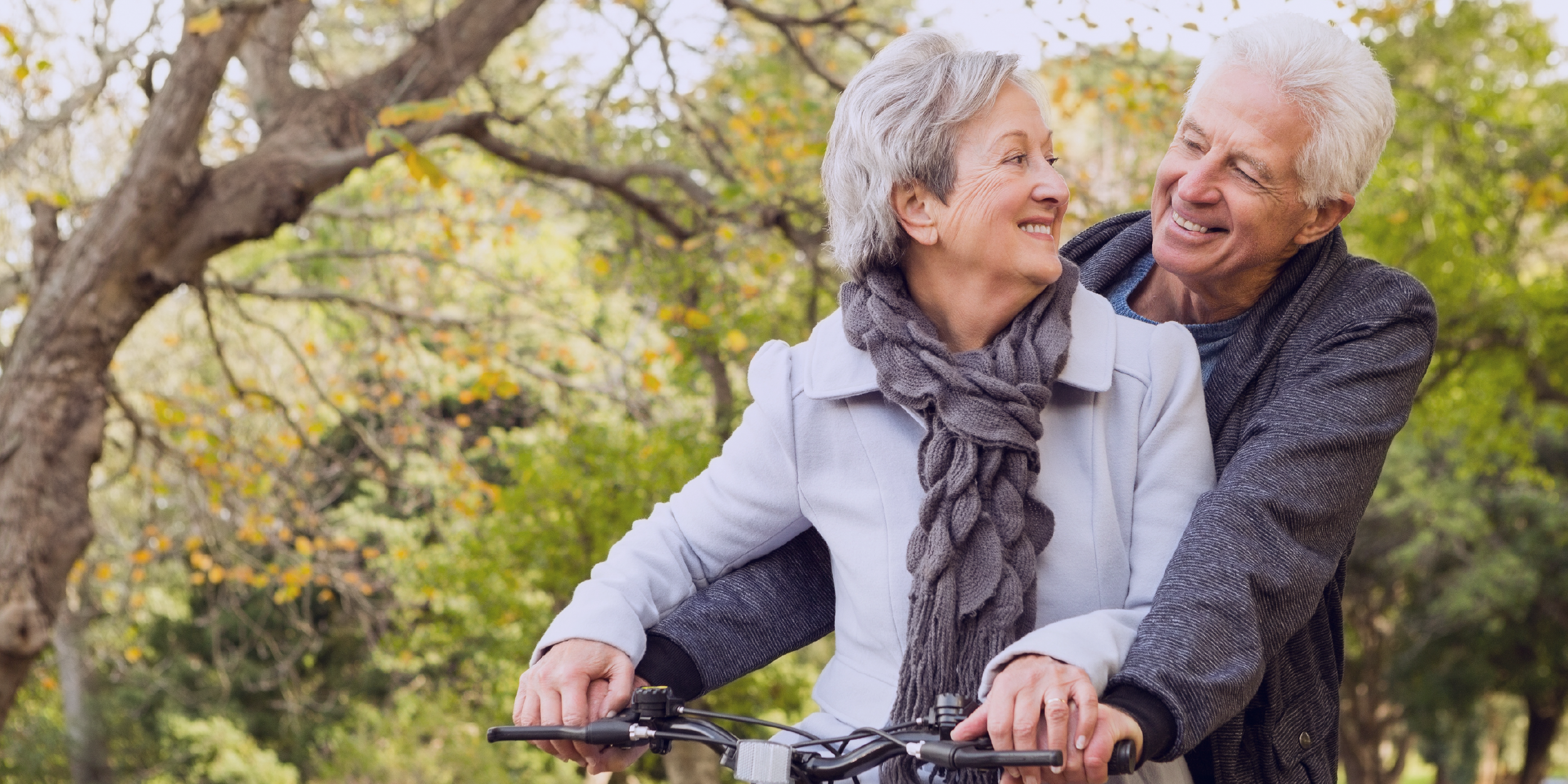 Foto de um belo casal idoso, com belos sorrisos, olhando um para o outro enquanto andam de bicicleta.